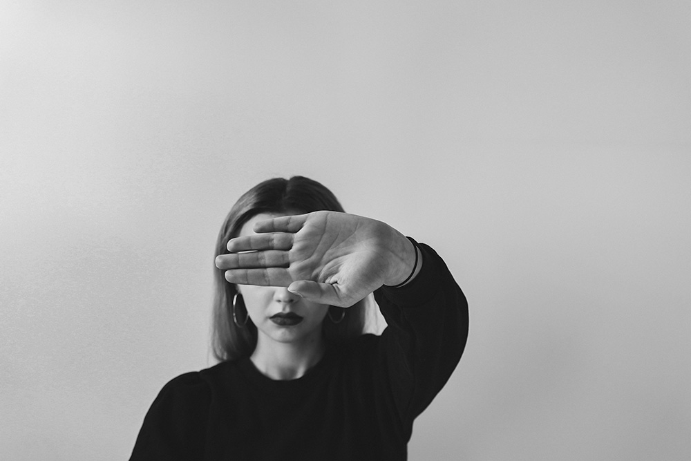 black and white photo of woman standing against a white wall with her hand up to the camera covering her eyes